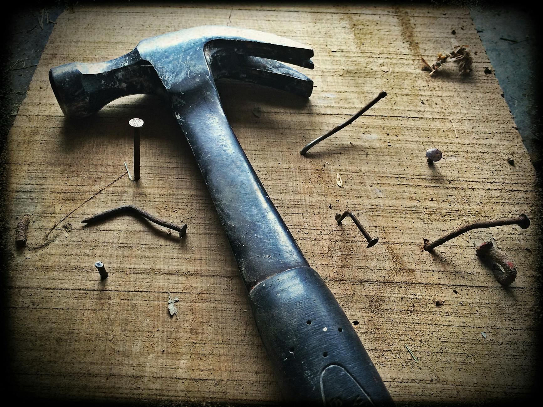 A hammer lying on a table surrounded by several nails. Some are bent, some are partly driven into the wood.